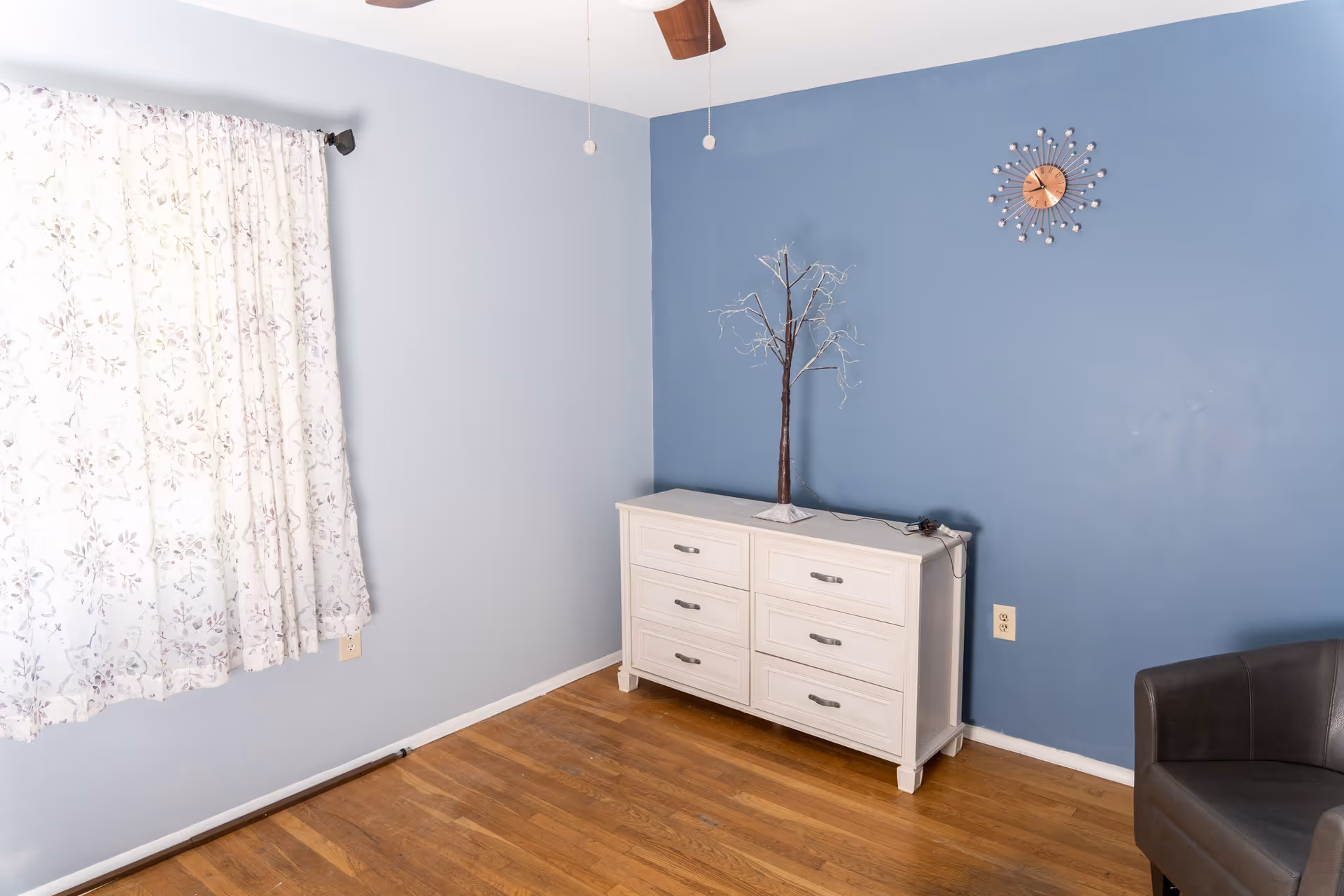 A room with light blue and darker blue walls, a wooden floor, a white dresser with six drawers, a decorative tree on top of the dresser, a modern wall clock, floral curtains covering a window, and a dark gray armchair partially visible on the right.