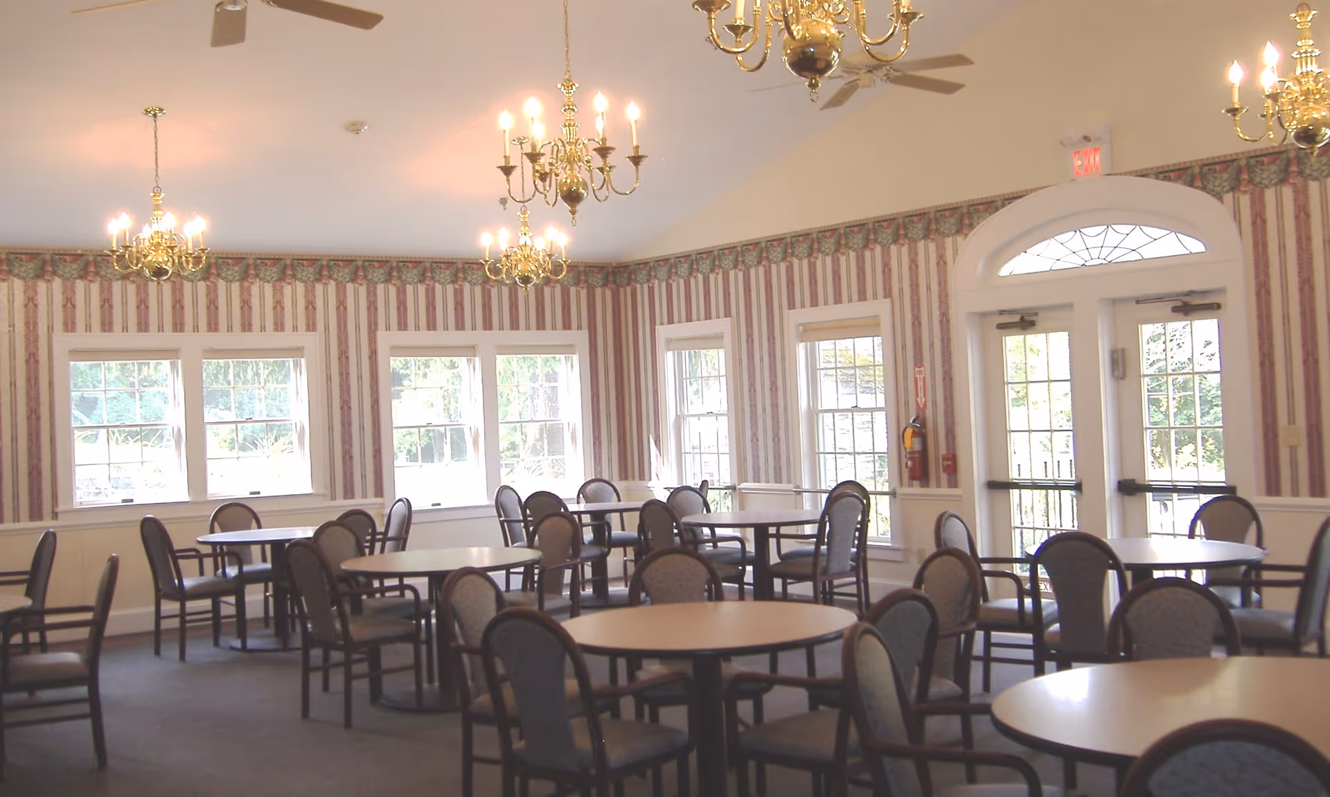 A dining room with multiple round tables and chairs arranged neatly. The room has patterned wallpaper with a floral border near the ceiling, several windows letting in natural light, and multiple brass chandeliers hanging from the ceiling. There is a double glass door with an arched window above it on one side of the room.