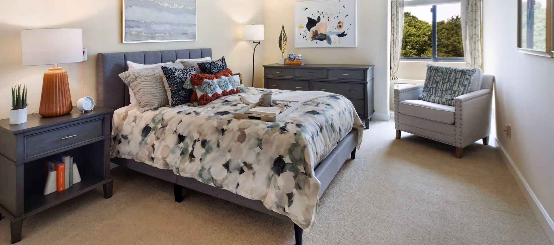 A well-lit bedroom with a bed featuring a floral-patterned comforter and multiple pillows. There is a gray nightstand with a lamp, clock, and a small plant on the left side of the bed. A gray dresser with decorative items and a framed abstract artwork is against the wall. A comfortable armchair with a patterned cushion is placed near a window showing green trees outside.
