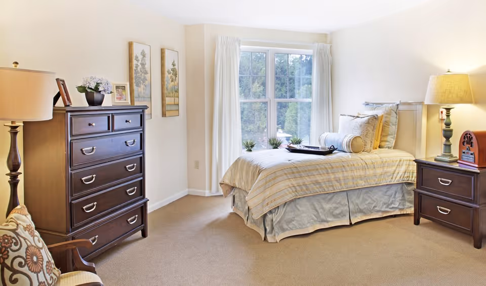 Sunny, well-lit bedroom with a single bed by a large window, wooden dresser and nightstand with lamps.