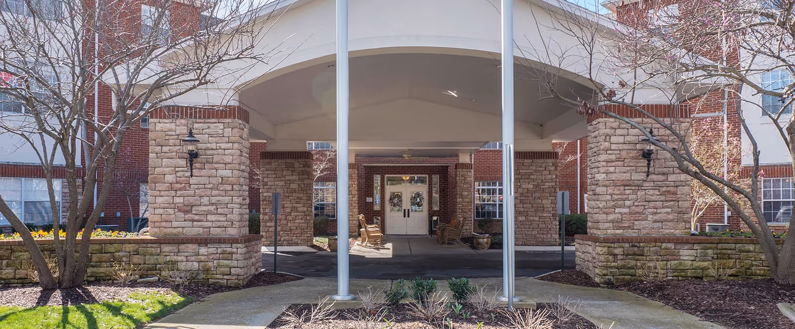 Entrance of a senior living facility with a covered driveway supported by stone pillars, brick walls, and a glass door entrance. There are leafless trees and landscaped areas with mulch and small plants on either side of the driveway.