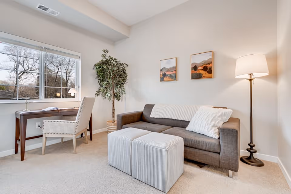 Bright living room with a gray sofa, two ottomans, floor lamp, desk by the window and a potted plant.