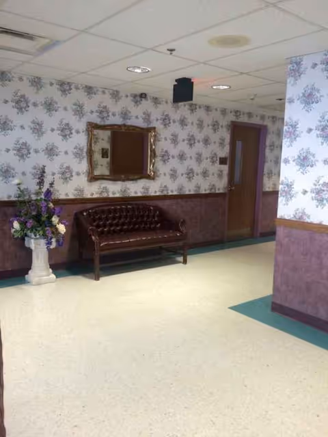 Leather sofa along a floral wallpapered nursing home hallway with a mirror and vase.