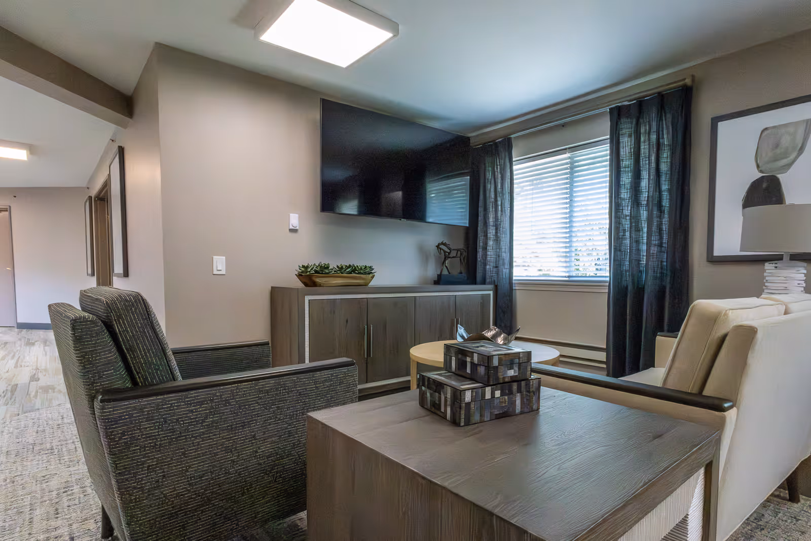 A modern living room area with two dark armchairs and a light-colored sofa arranged around a wooden coffee table and a wooden cabinet. A flat-screen TV is mounted on the wall above the cabinet. A window with blinds and dark curtains lets in natural light. The room has neutral-colored walls and carpeted flooring.