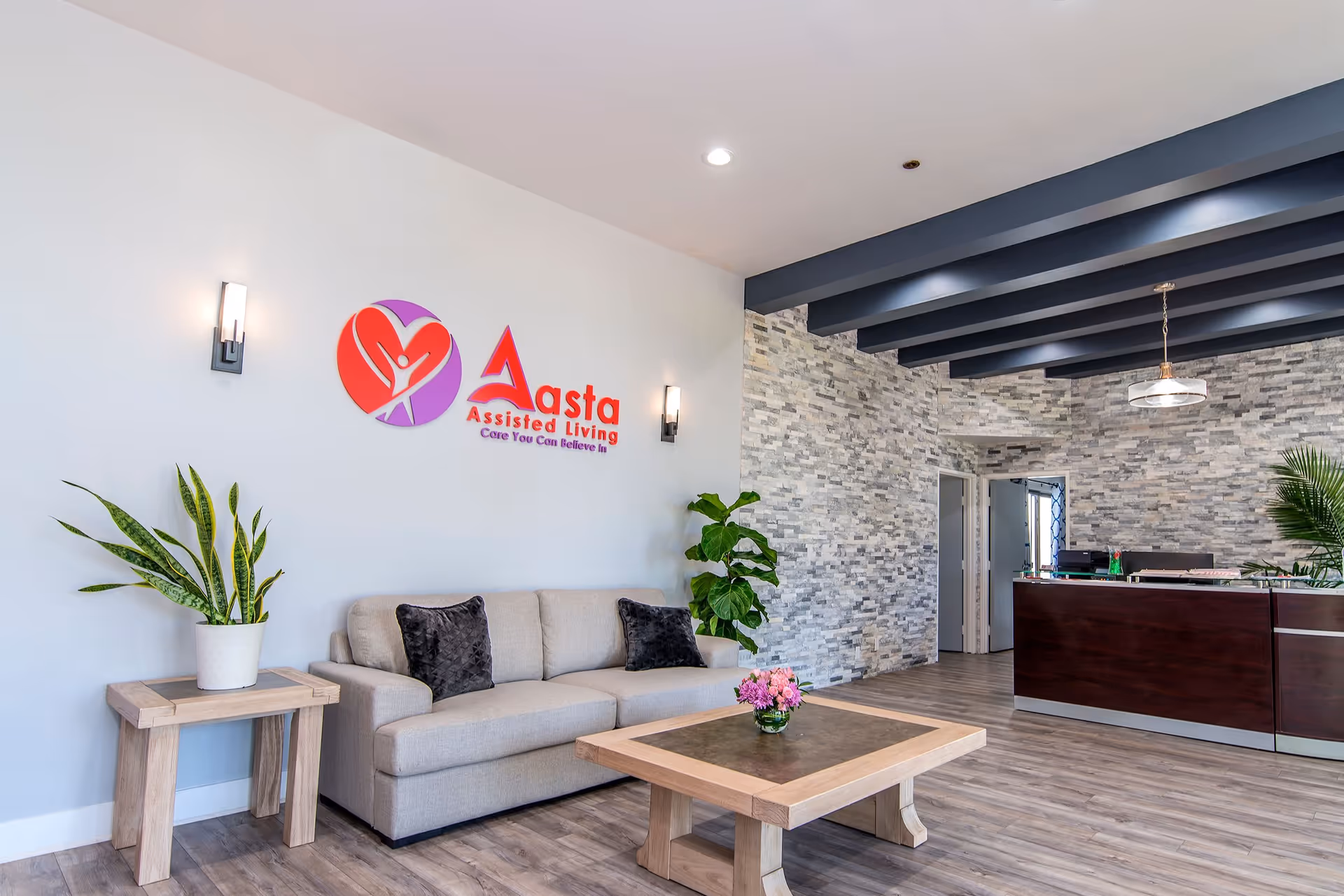 A modern assisted living facility lobby with a beige sofa, two black pillows, a wooden coffee table with a small flower arrangement, a side table with a potted plant, and a reception desk in the background. The walls are decorated with a stone-like texture and a logo for Aasta Assisted Living is displayed on a white wall.