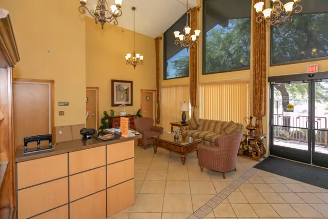 Interior view of a senior living facility lobby with a reception desk on the left, a seating area with a sofa and two armchairs around a wooden coffee table, large windows with vertical blinds, and chandeliers hanging from a high ceiling. There is a glass exit door on the right side with a view of trees outside.