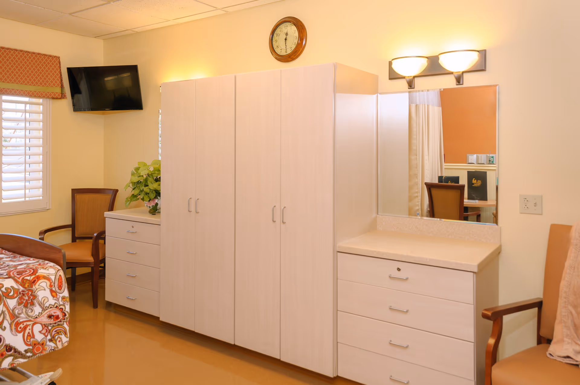 A well-lit senior living bedroom featuring a bed with a colorful patterned bedspread, a wooden chair with armrests, a wall-mounted flat-screen TV, a large wardrobe with multiple doors, a dresser with drawers, a countertop with a mirror above it, and a wall clock. The room has light-colored walls and flooring.