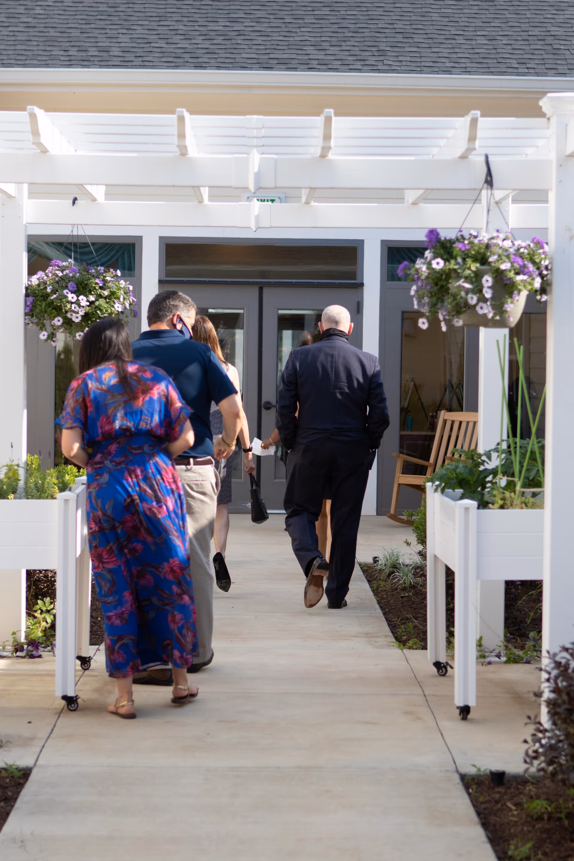 Several people walking toward the entrance of a senior living building beneath a white pergola with hanging flower baskets and planter boxes.