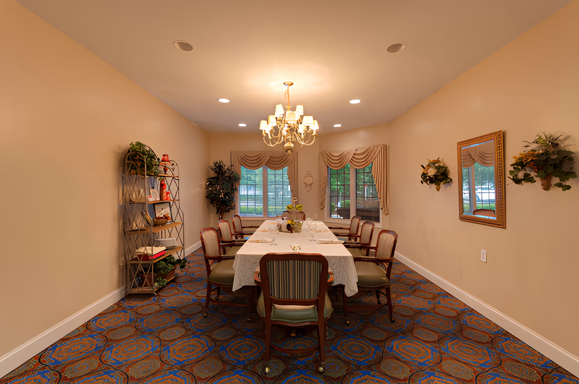 A formal dining room with a long rectangular table covered with a white tablecloth, surrounded by eight wooden chairs with cushioned seats. The room has beige walls, a patterned carpet with blue and brown tones, and a chandelier hanging from the ceiling. There are two large windows with beige drapes at the far end, a decorative shelf with plants and items on the left wall, and a mirror with floral wall decorations on the right wall.