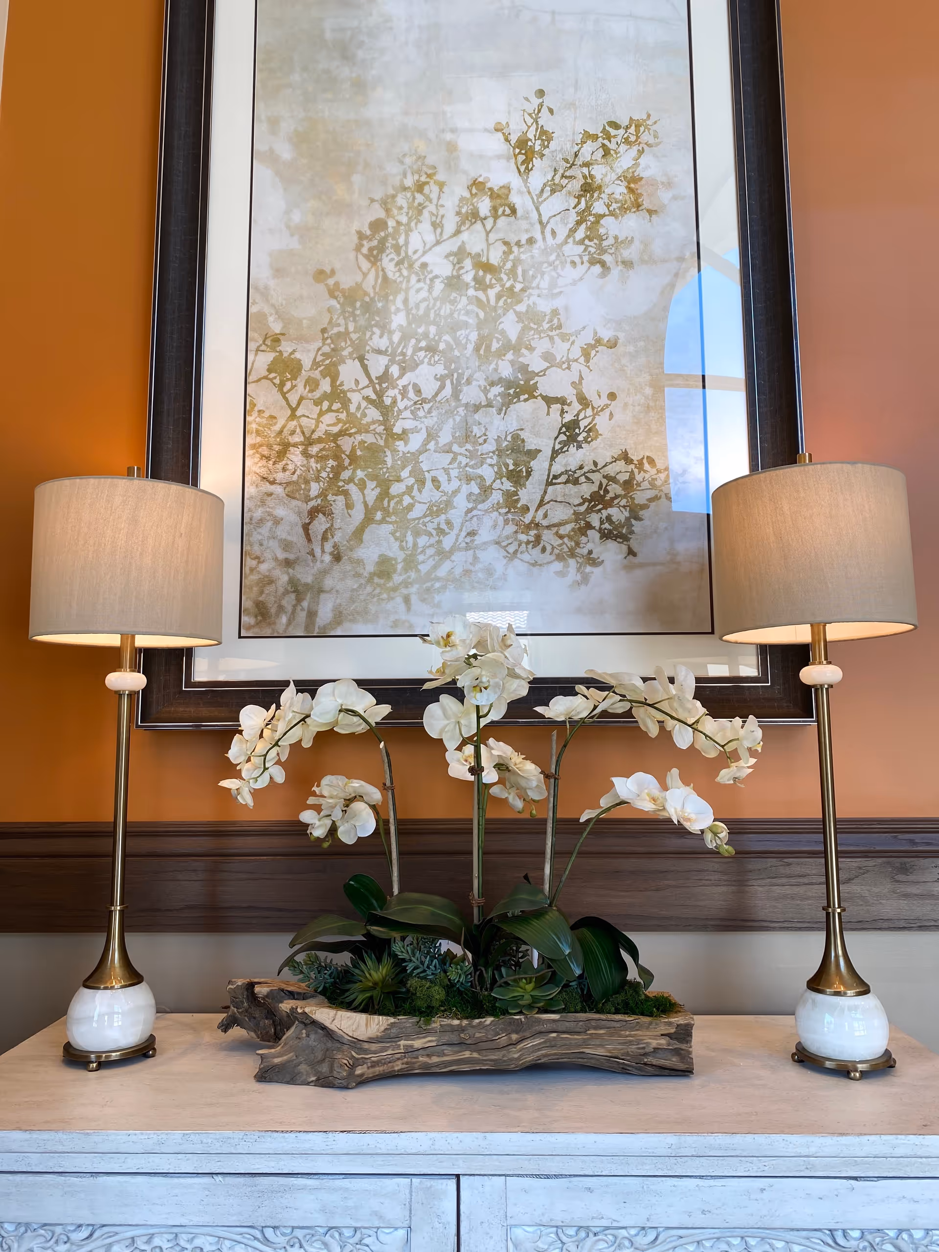 A decorative arrangement on a white wooden console table featuring two tall table lamps with beige shades and brass and white bases. Between the lamps is a wooden planter with white orchid flowers and green foliage. Behind the arrangement is a large framed artwork depicting a subtle botanical design on a beige background, mounted on a warm orange wall.