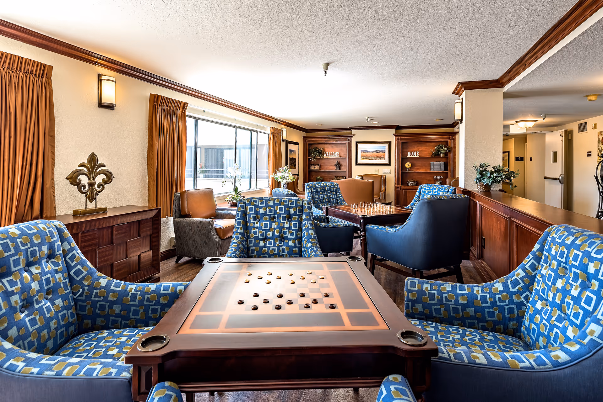 A cozy senior living common area with multiple seating arrangements including blue patterned armchairs and brown leather chairs. There are two wooden game tables with checkers and chess pieces set up. The room has large windows with brown curtains, wooden cabinetry, and decorative items on shelves. The lighting is warm and inviting.