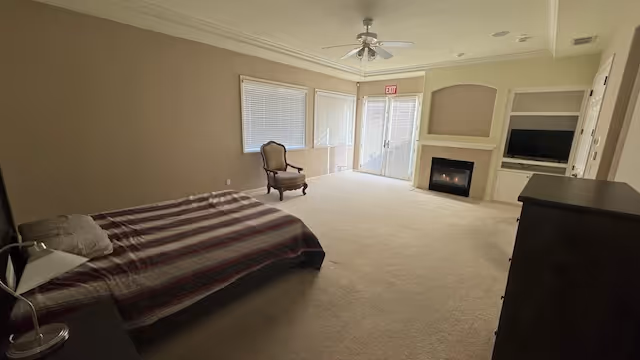 A spacious bedroom with a single bed covered with a striped blanket, a bedside lamp, an armchair near large windows, a fireplace, a built-in shelving unit with a TV, and a dresser. The room has beige walls and carpeted floor, with a ceiling fan and glass doors leading outside.