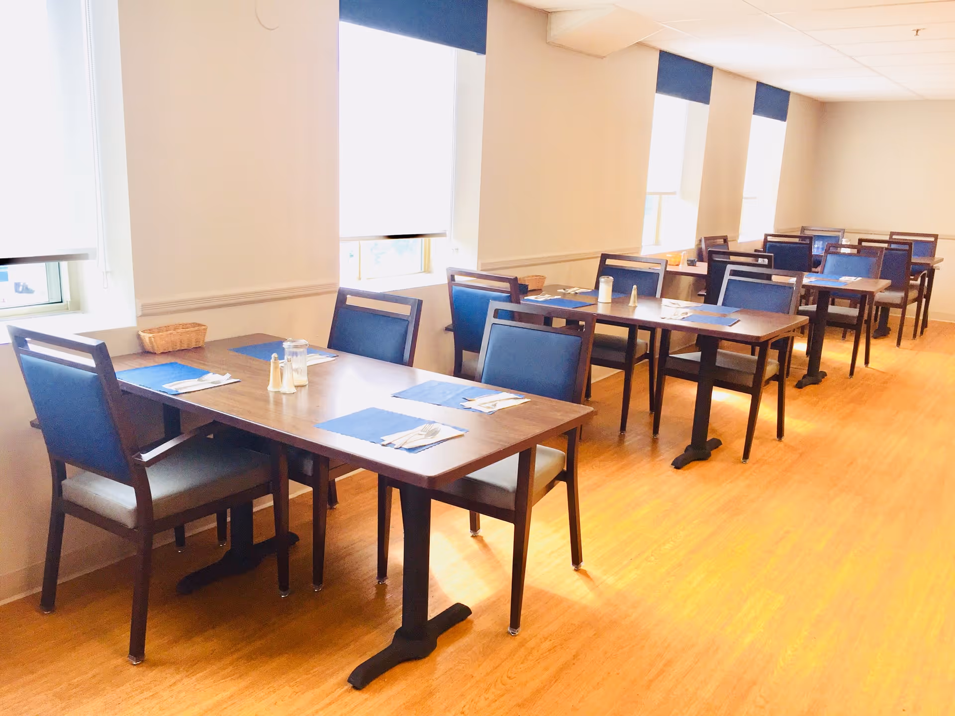 A bright dining room with several wooden tables and blue cushioned chairs arranged neatly. Each table has blue placemats, silverware wrapped in napkins, and salt and pepper shakers. Large windows with white blinds allow natural light to fill the room, which has light-colored walls and a wooden floor.
