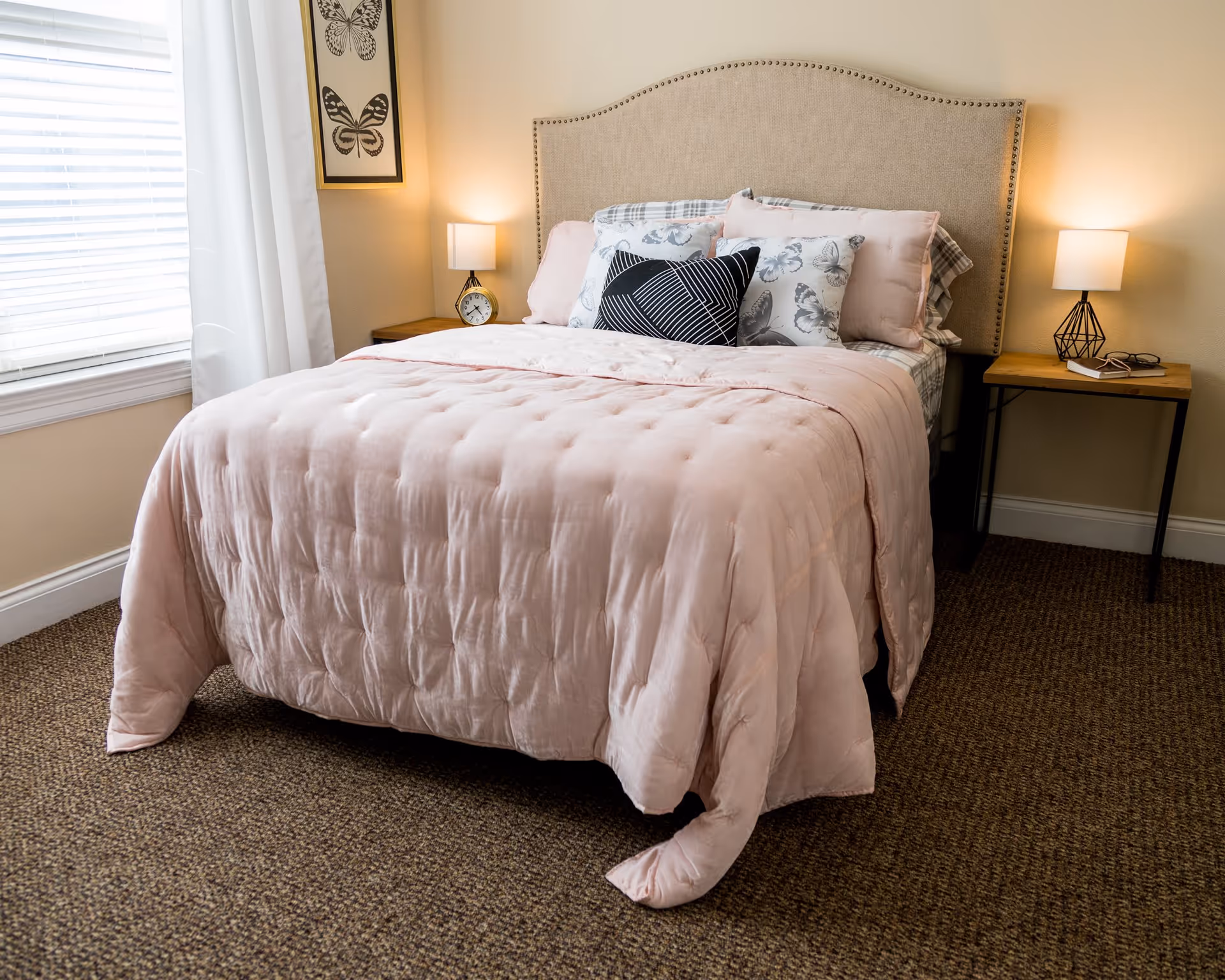 A neatly made bed with a beige upholstered headboard, pink comforter, and multiple pillows including patterned and solid colors. There are two wooden nightstands on either side of the bed, each with a lamp. A window with white curtains is on the left side, and a framed butterfly artwork hangs on the wall above the left nightstand.