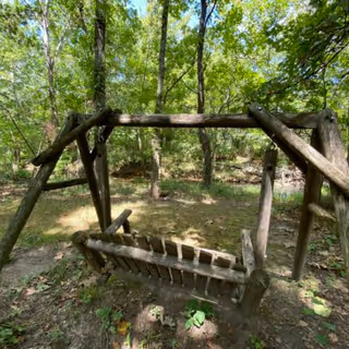 A rustic wooden swing set made from logs situated in a wooded outdoor area with green trees and grass surrounding it.
