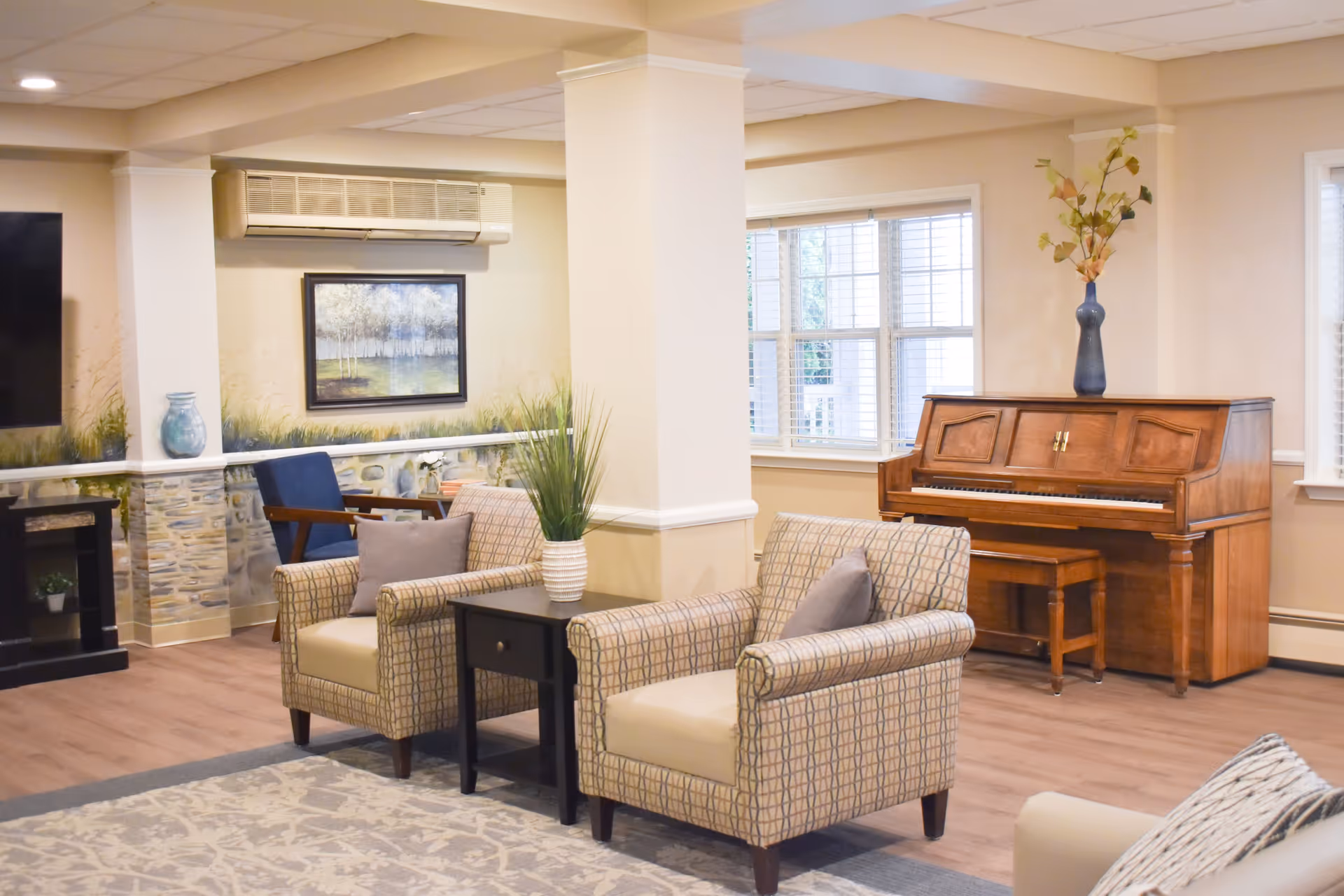A cozy senior living room with two patterned armchairs separated by a small black side table with a white vase holding green plants. In the background, there is a wooden upright piano with a blue vase and decorative branches on top. The room has large windows with white blinds, a wall-mounted air conditioning unit, a framed landscape painting, and a blue armchair near the corner. The floor is wood with a patterned area rug.