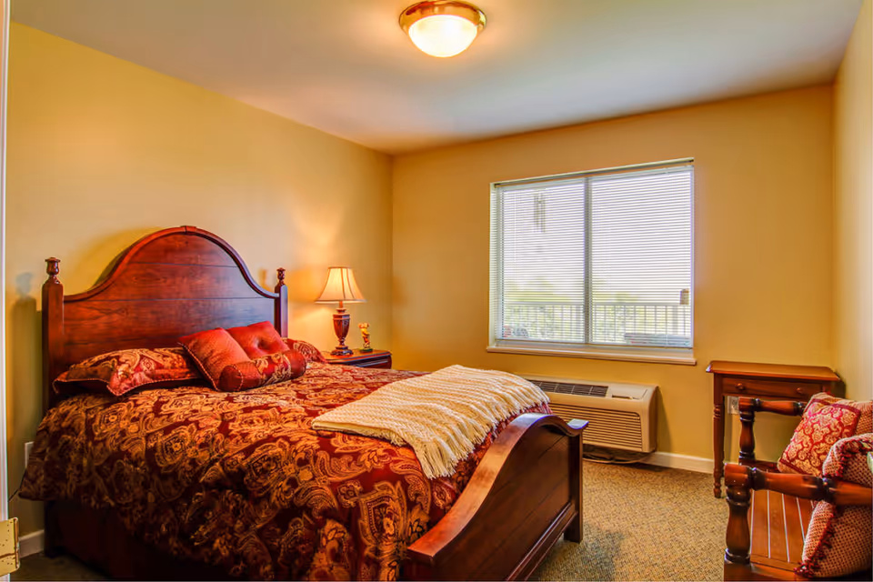 A cozy bedroom with a wooden bed frame and headboard, covered with a patterned red and gold bedspread and matching pillows. A bedside table with a lamp is next to the bed. There is a window with blinds letting in natural light, and a heating/cooling unit below the window. A wooden chair with cushions and a small wooden table are also visible in the room.