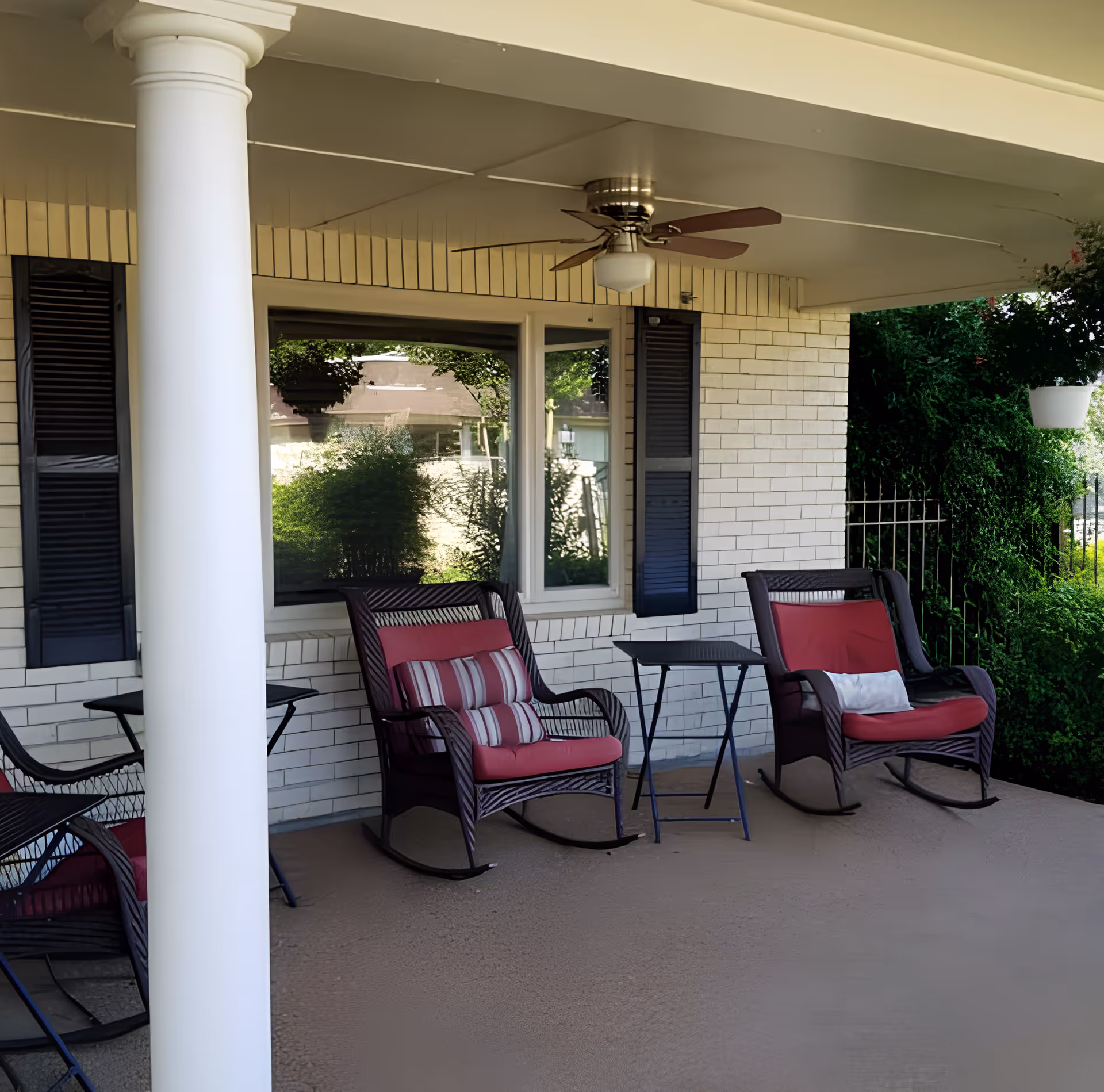 Covered outdoor patio area with two cushioned rocking chairs and a small table between them. The patio has a ceiling fan, white brick walls with black shutters, and greenery visible in the background.