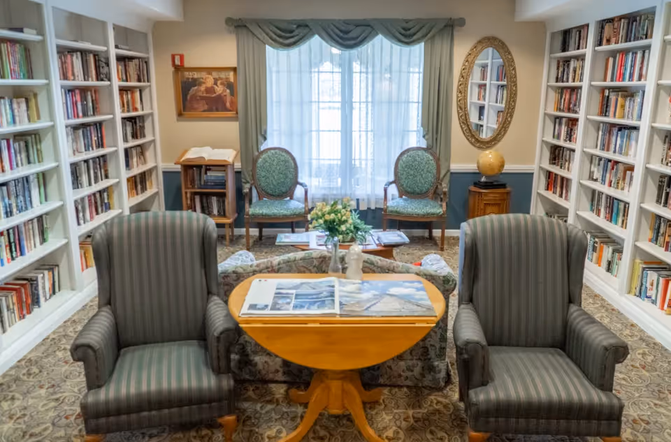 Cozy library-style sitting room with bookshelves on both sides, two striped armchairs facing a round wooden table and a window with two upholstered chairs.