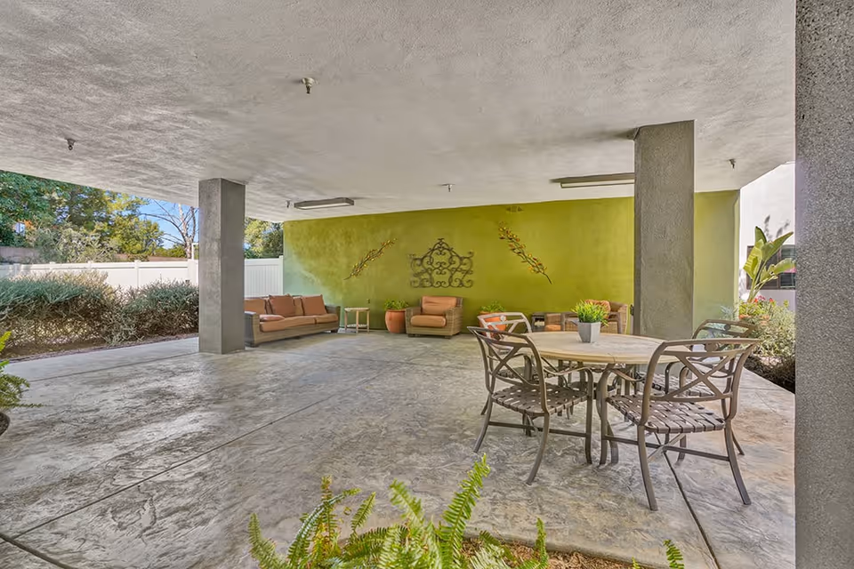 Covered outdoor seating area with a round table and four metal chairs in the foreground, and cushioned sofas and armchairs against a green wall decorated with metal wall art in the background. The area is supported by concrete pillars and surrounded by plants and bushes.
