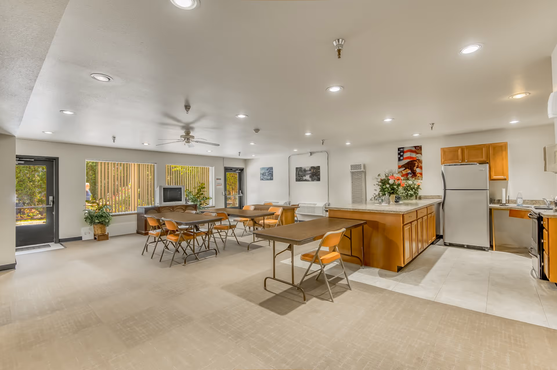 A spacious common area in a senior living facility featuring a kitchen with wooden cabinets, a refrigerator, and a stove. There are several tables with orange chairs arranged for dining or activities. The room has large windows letting in natural light, potted plants, and a ceiling fan. The walls are decorated with framed pictures and a floral arrangement is placed on the kitchen island.