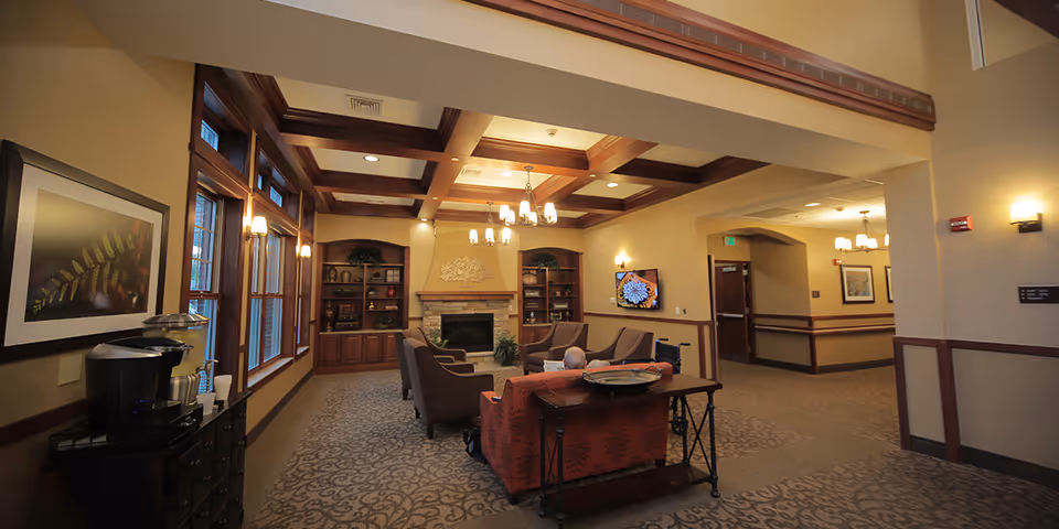 A cozy senior living facility common area with a fireplace, wooden ceiling beams, and built-in bookshelves. The room is furnished with comfortable armchairs and a sofa arranged around a coffee table. Large windows line one wall, letting in natural light. A coffee station is visible on the left side, and a flat-screen TV is mounted on the wall to the right. Warm lighting fixtures and neutral tones create a welcoming atmosphere.