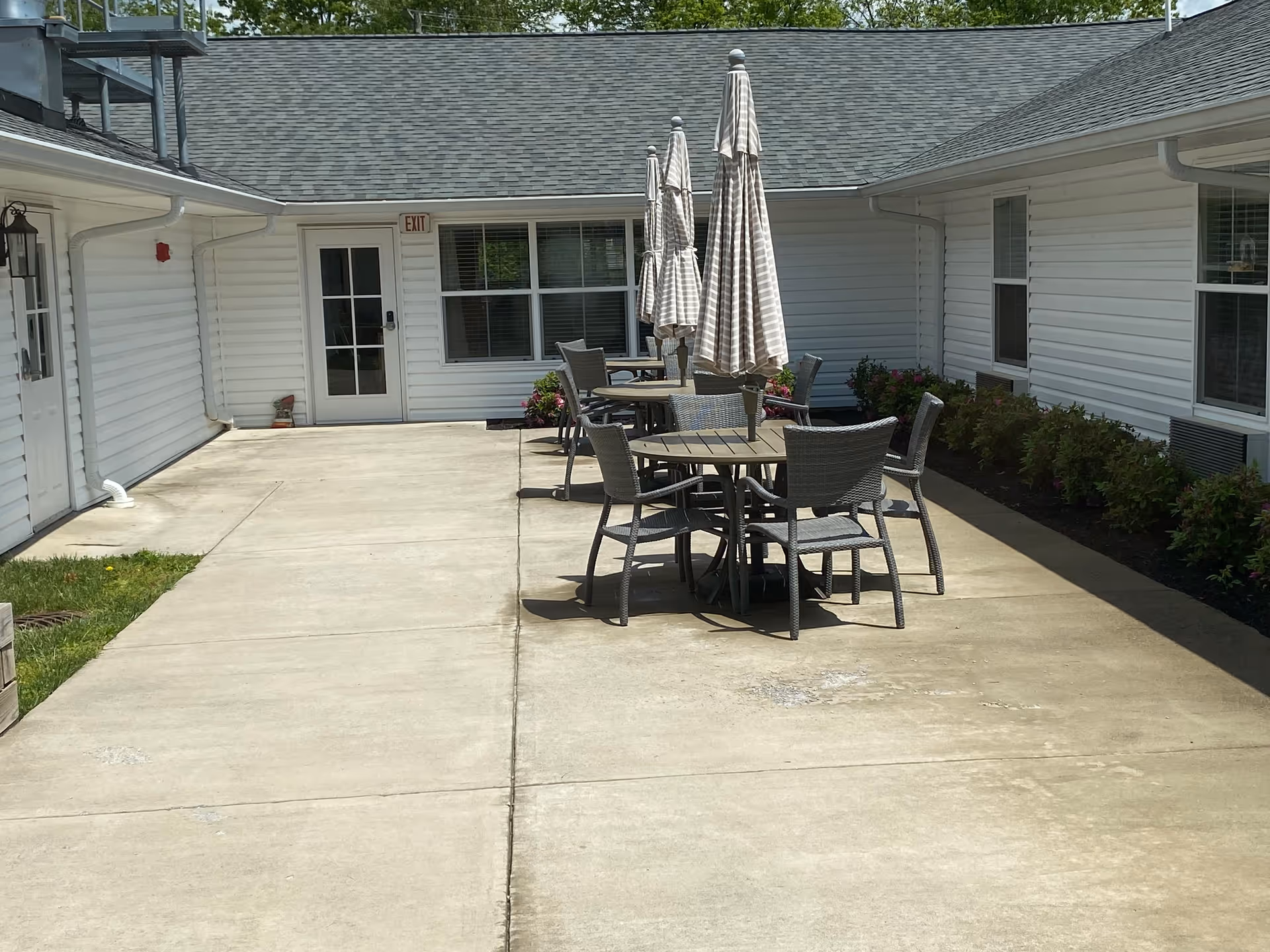 Outdoor patio area at Greenfield Reflections of Woodstock with round tables and chairs arranged along a concrete walkway. Each table has a closed striped umbrella. The patio is surrounded by white siding buildings and some greenery along the edges.