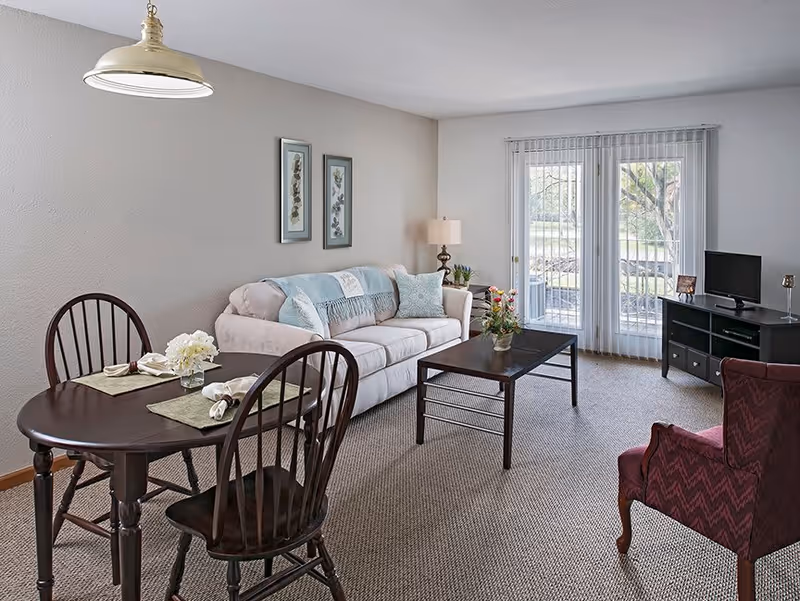 A cozy senior living room with a beige sofa adorned with blue and patterned pillows, a dark wooden coffee table with a flower vase, a matching dining table set with two chairs and place settings, a burgundy armchair, a TV on a black stand, and large glass doors with vertical blinds leading outside.