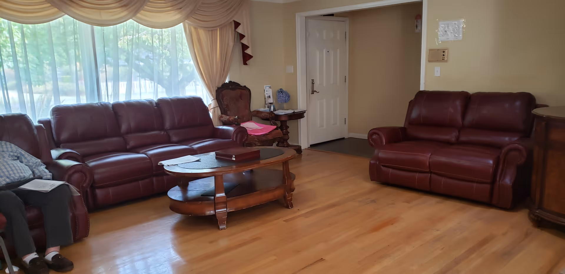 A living room with two brown leather sofas, a wooden coffee table in the center, a wooden side table with a chair in the corner, and large windows with sheer curtains allowing natural light to fill the room. The floor is wooden, and there is a white door in the background.
