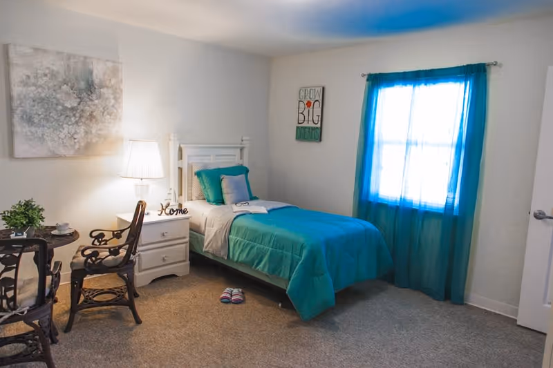 A cozy bedroom with a single bed made up with teal bedding and pillows. Next to the bed is a white nightstand with a lamp and a decorative 'Home' sign. There is a small round table with two chairs on the left side, and a window with teal curtains on the right wall. A framed artwork and a motivational sign that reads 'Grow Big Dreams' are hung on the walls.