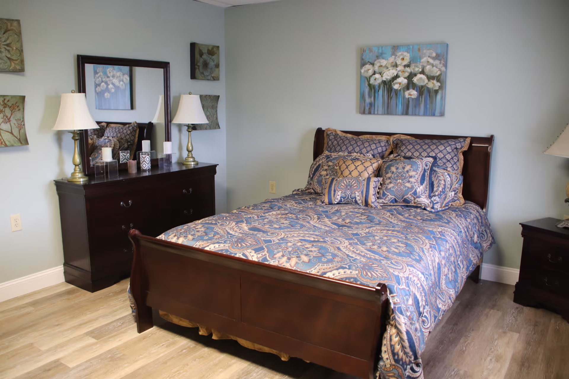A bedroom with a wooden bed frame and a blue patterned bedspread with multiple pillows. There is a dark wooden dresser with a large mirror above it, two table lamps, and decorative items on top. The walls are light blue with floral artwork hanging on them. The floor is light wood.