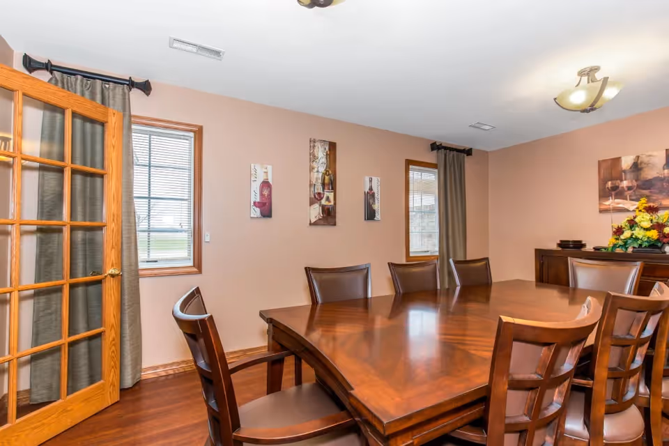A dining room with a large polished wooden table surrounded by eight matching wooden chairs with leather seats. The room has light brown walls adorned with wine-themed artwork. Two windows with blinds and dark curtains flank the walls. A wooden sideboard with a floral arrangement and plates is visible in the background. A wooden door with glass panes is partially open on the left side.