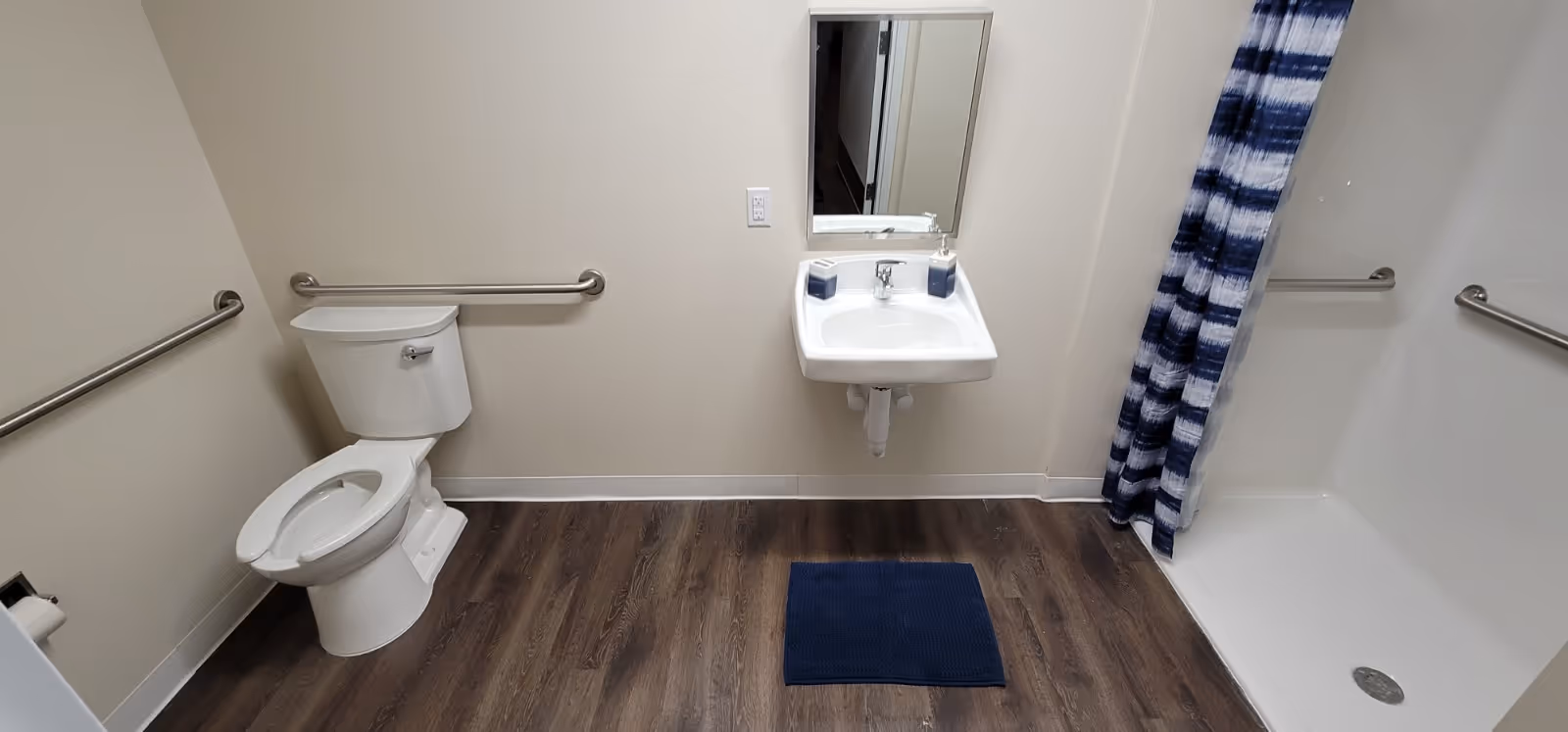 A clean bathroom with a white toilet, a wall-mounted sink with a mirror above it, and a shower area with a blue and white striped curtain. The floor is wood-style, and there are metal grab bars on the walls near the toilet and shower for accessibility.