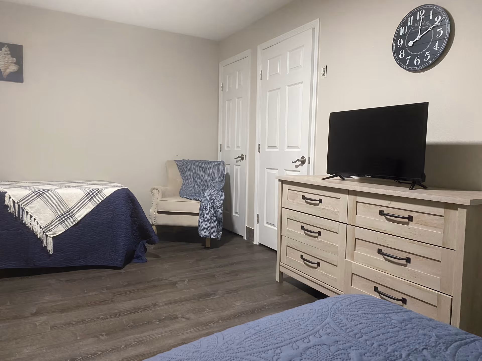 A senior living facility bedroom with two beds covered in blue and white blankets, a beige armchair with a blue throw draped over it, a light wood dresser with six drawers, a flat-screen TV on top of the dresser, and a wall clock above the TV. The room has light-colored walls and wood flooring.