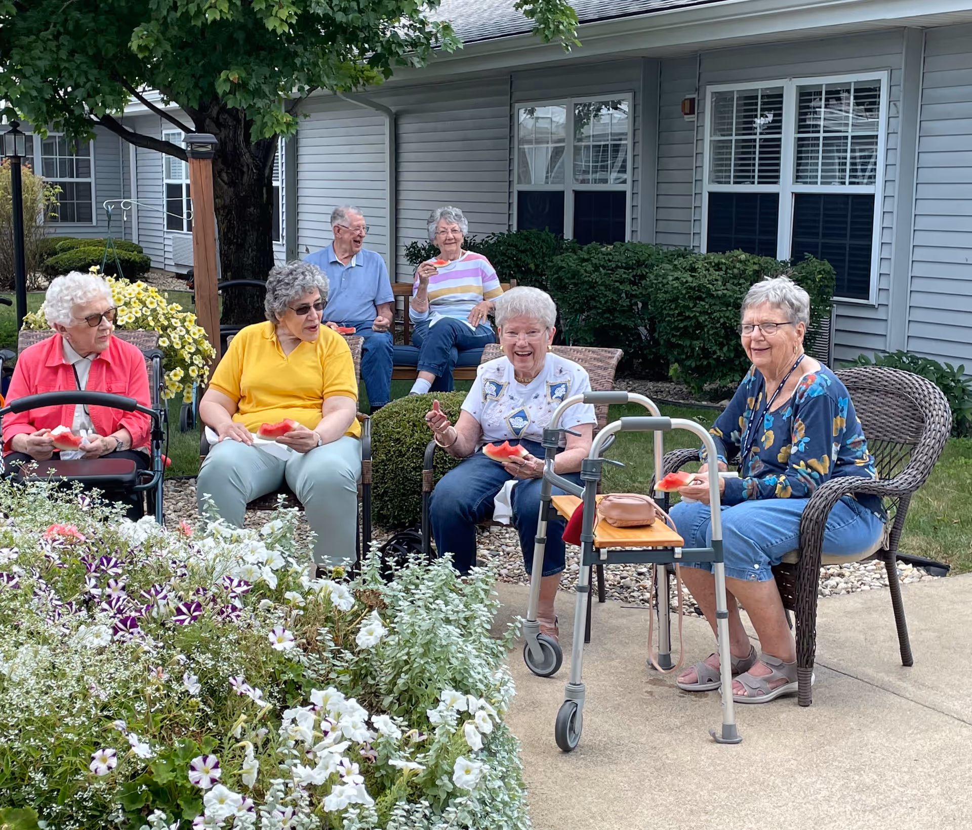 A group of elderly people sitting outdoors in a garden area of a senior living facility. They are seated on chairs and benches near a building with gray siding and white-trimmed windows. Some are holding slices of watermelon and appear to be enjoying a social gathering. There are flowers and greenery in the foreground and background.
