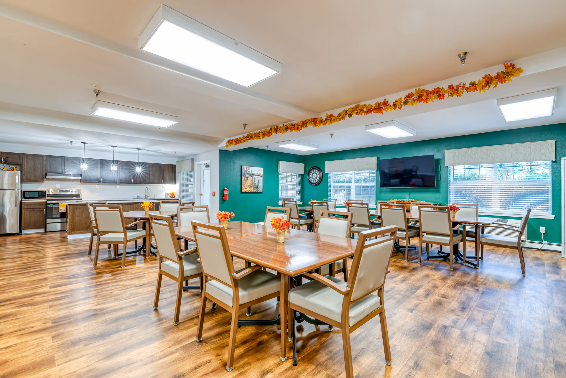 A bright and spacious dining room with multiple wooden tables and cushioned chairs arranged neatly. The room features a kitchen area with stainless steel appliances including a refrigerator, stove, and microwave. The walls are painted green with large windows allowing natural light to enter. There is a wall-mounted TV and a large clock on the wall. Autumn-themed garland decorates the ceiling.