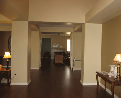 Interior view of a senior living facility showing a hallway with wooden flooring leading to a living area with chairs and a fireplace. There are two side tables with lamps on either side of the hallway.