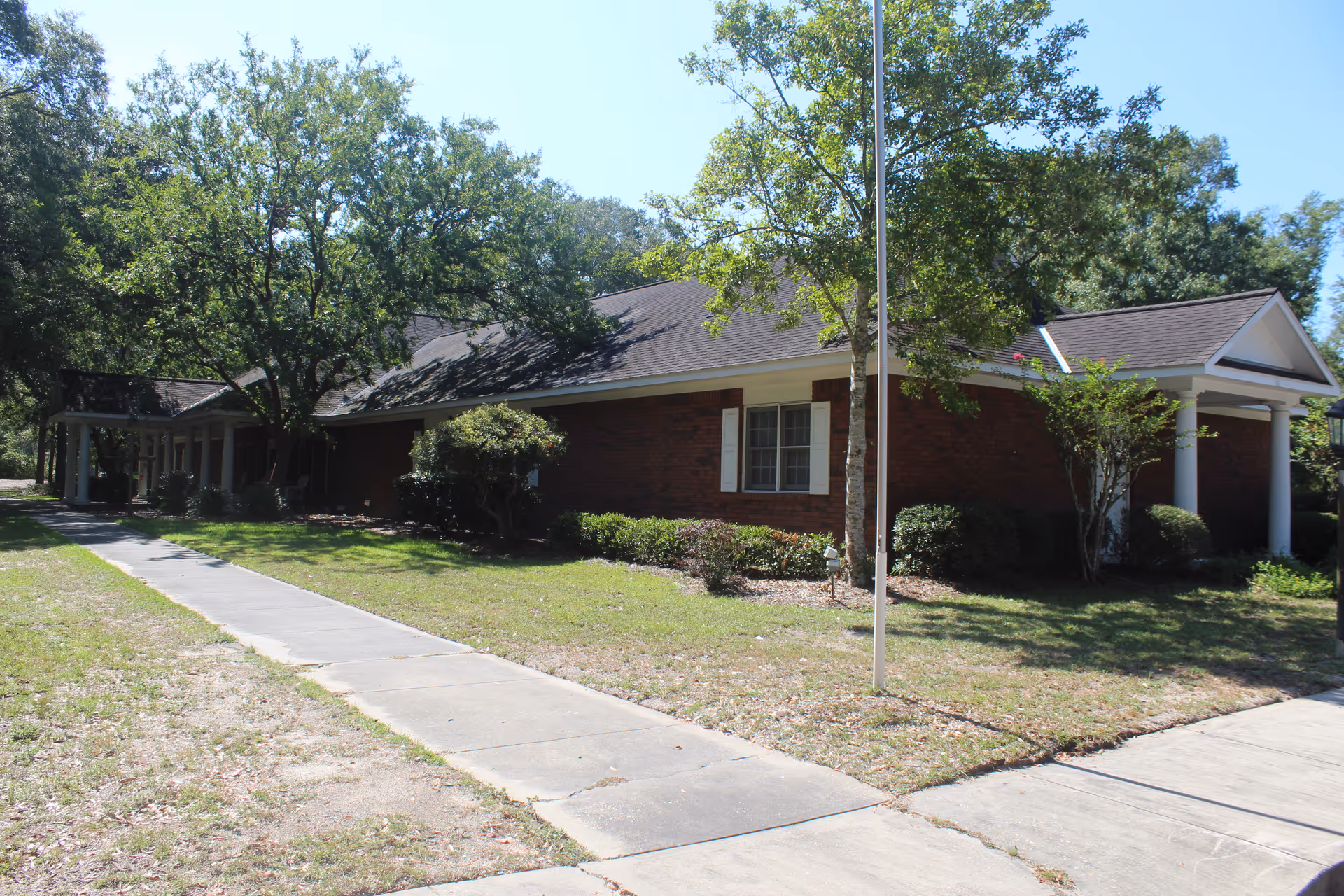 Single-story brick assisted living building with a sidewalk, trees, and a flagpole in front.