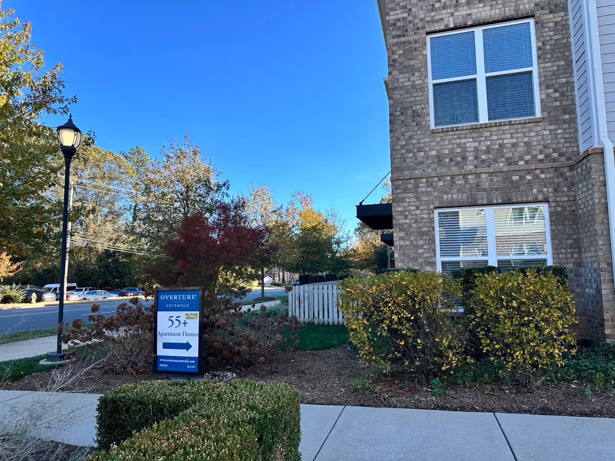 Exterior front of a brick apartment building with landscaping and a sign reading 'Overture Cotswold 55+ Apartment Homes'.