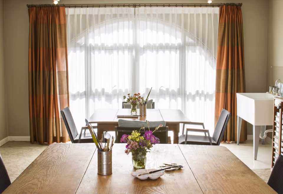 A bright room with large arched windows covered by sheer white curtains and orange striped drapes. There are wooden tables with chairs around them, decorated with small vases of flowers and containers holding paintbrushes and art supplies. A white utility sink is visible on the right side of the room.