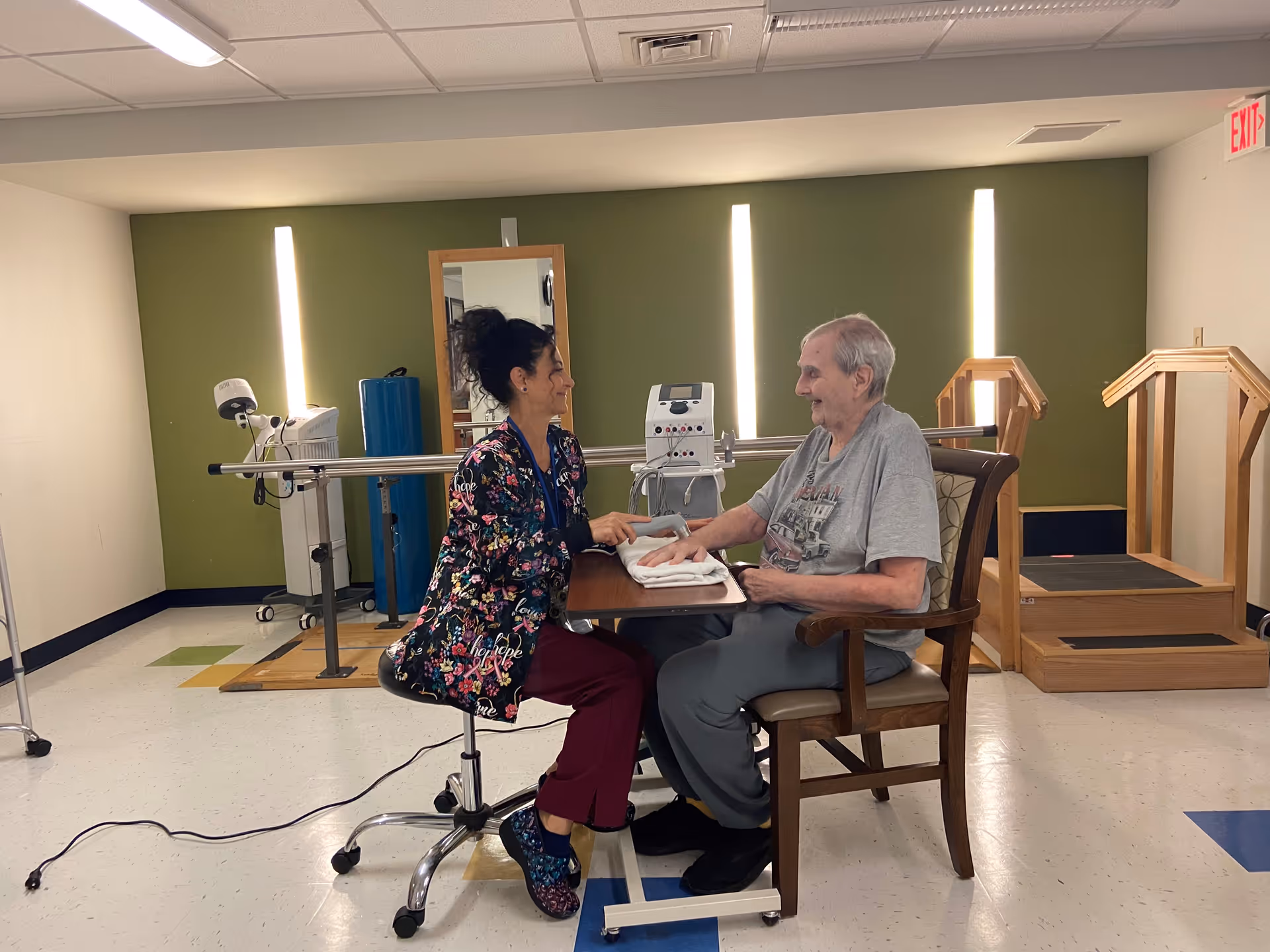 A healthcare worker in colorful scrubs sitting on a rolling stool, smiling and holding a device to the hand of an elderly man seated across from her at a small table in a rehabilitation room with exercise equipment and parallel bars in the background.