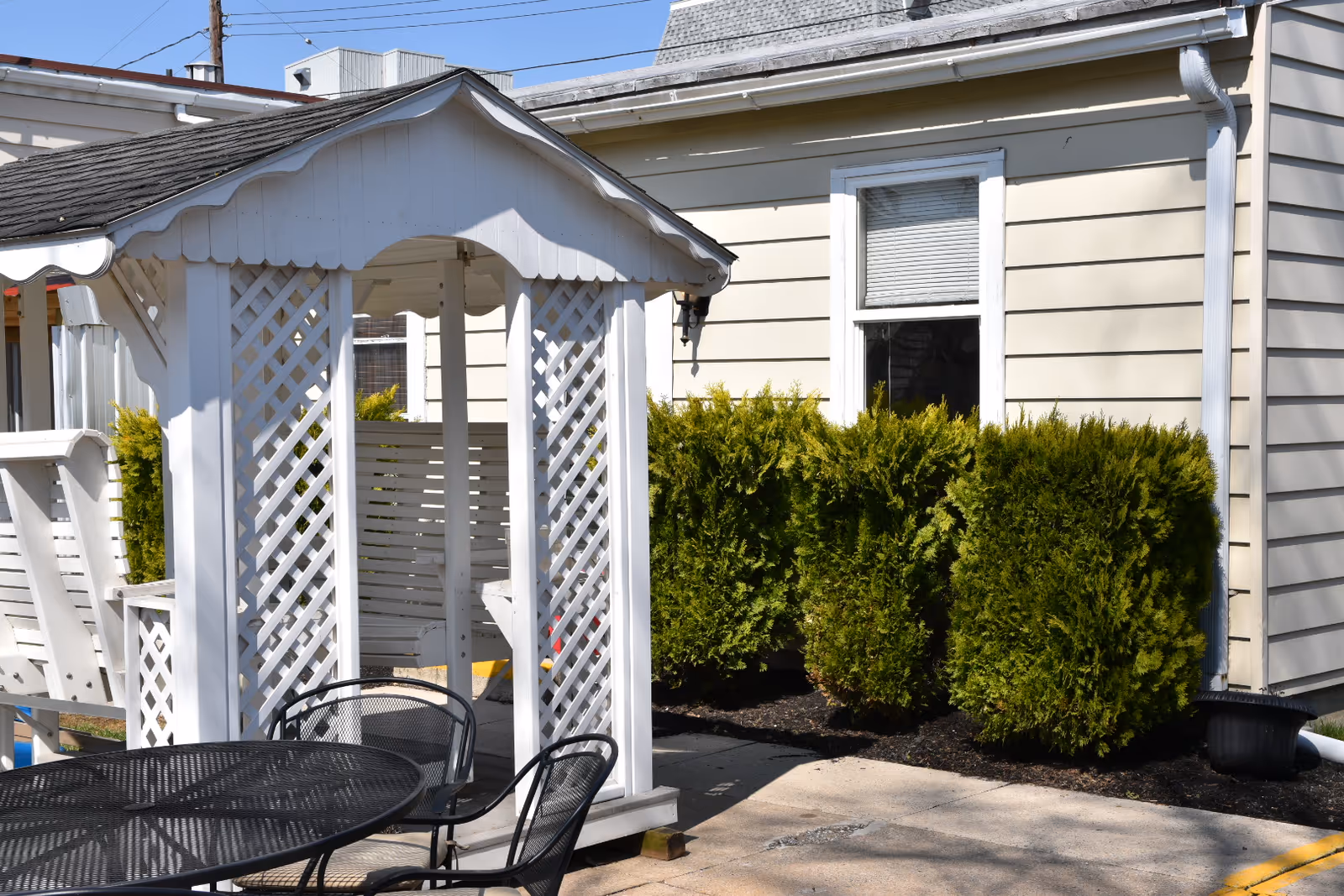 Outdoor seating area with a black metal table and chairs next to a white wooden gazebo structure with lattice sides. Behind the gazebo is a beige building with a window and green bushes along the wall.