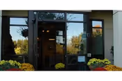 Glass entrance doors to a building with reflections of trees and sky, flanked by large windows and flower pots with yellow and orange flowers on either side.