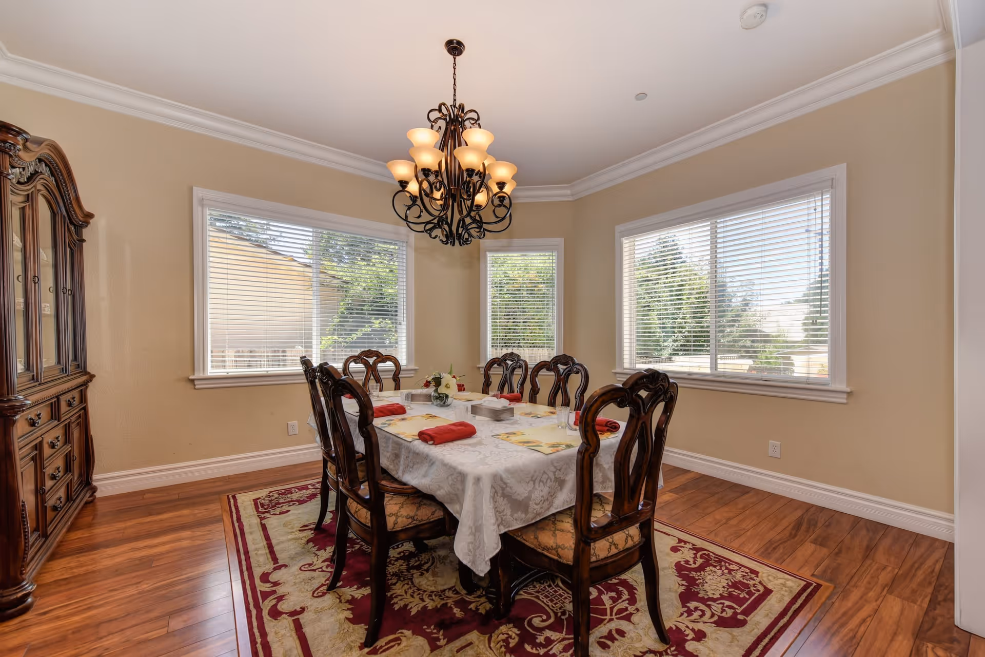 A dining room with a wooden dining table covered with a white tablecloth and set with placemats and red napkins. Six ornate wooden chairs surround the table. A decorative chandelier hangs above the table. The room has beige walls, three large windows with white blinds, a wooden floor, and a red patterned area rug. A wooden china cabinet is visible on the left side of the room.