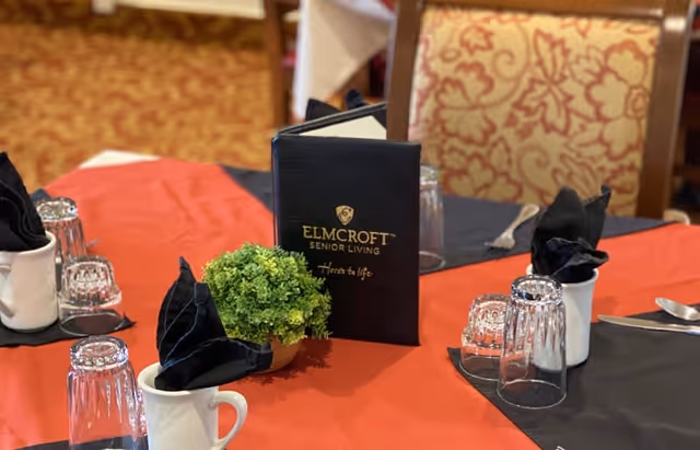 A dining table set with a red and black tablecloth, white mugs with black folded napkins, upside-down drinking glasses, silverware, a small green plant centerpiece, and a black menu card with the text 'ELMCROFT SENIOR LIVING Heart to life'. The background shows patterned upholstered chairs.