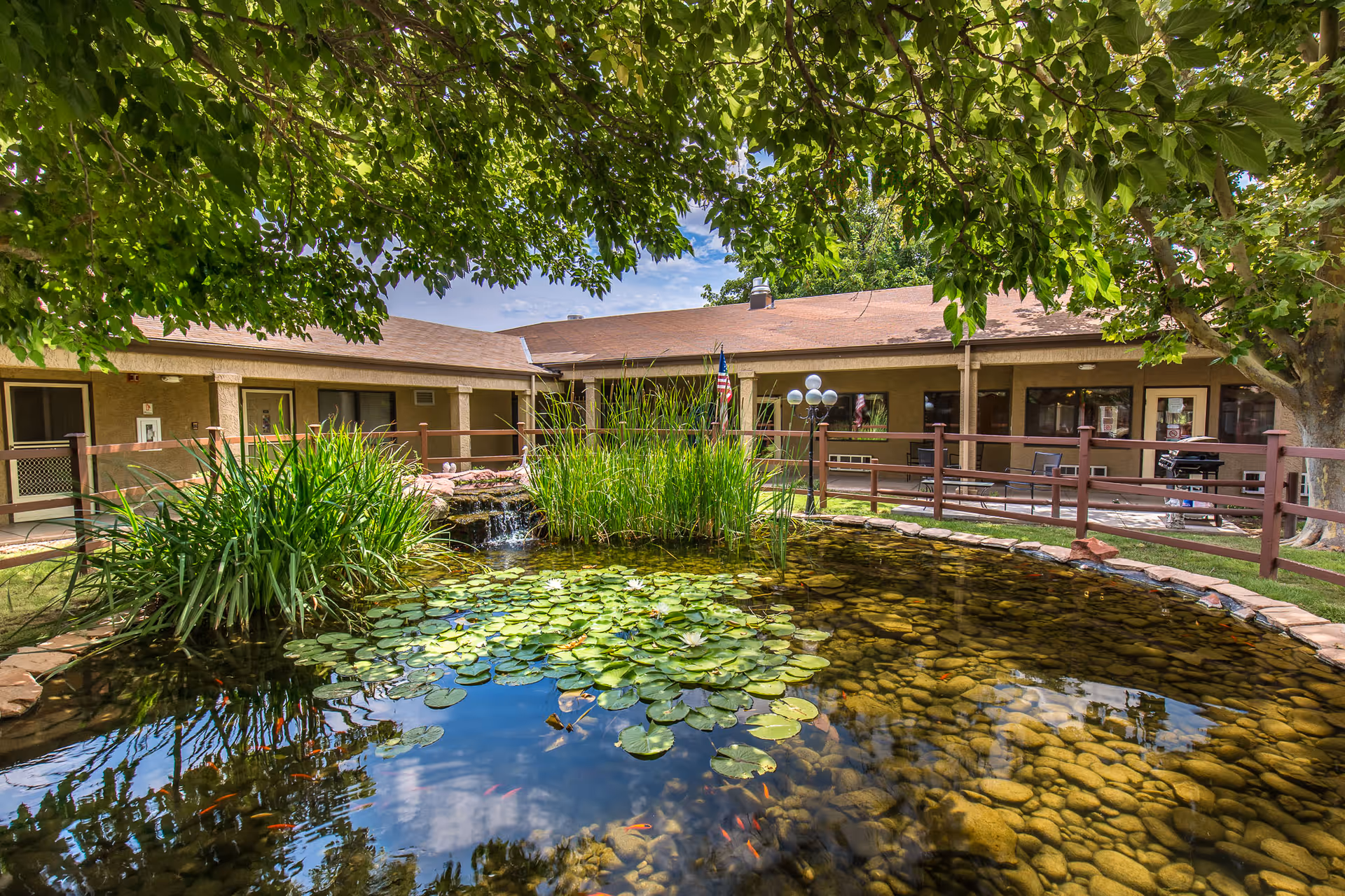 A tranquil outdoor courtyard at Verde Valley Assisted Living featuring a pond with lily pads and small fish, surrounded by green plants and trees. The courtyard is enclosed by a building with a covered walkway and several windows and doors. There is a wooden railing around the pond and a lamp post with multiple globes near the center.
