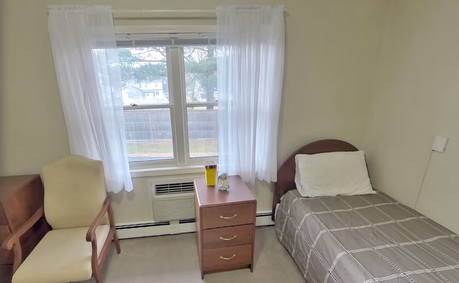 A small bedroom with a single bed covered with a gray patterned blanket and a white pillow. Next to the bed is a wooden nightstand with three drawers, holding a yellow container and a small decorative item. To the left of the nightstand is a beige upholstered armchair with wooden armrests. A window with sheer white curtains lets in natural light, and below the window is a wall-mounted air conditioning unit.