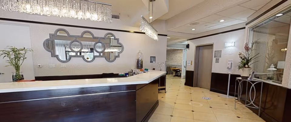 Reception area with a dark wood and white marble counter, a decorative mirror on the wall, hanging crystal light fixtures, and a hallway leading to an elevator and a dining area in the background. There are plants and decorative items on the counter and side table.