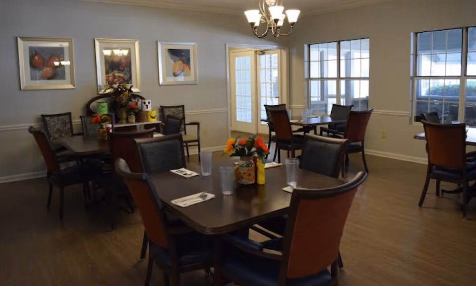 A dining room with several dark wooden tables and chairs arranged neatly. Each table has a small floral centerpiece and place settings with napkins, utensils, and plastic cups. The room has light-colored walls adorned with framed artwork and a chandelier hanging from the ceiling. Large windows and glass doors allow natural light to enter the space.