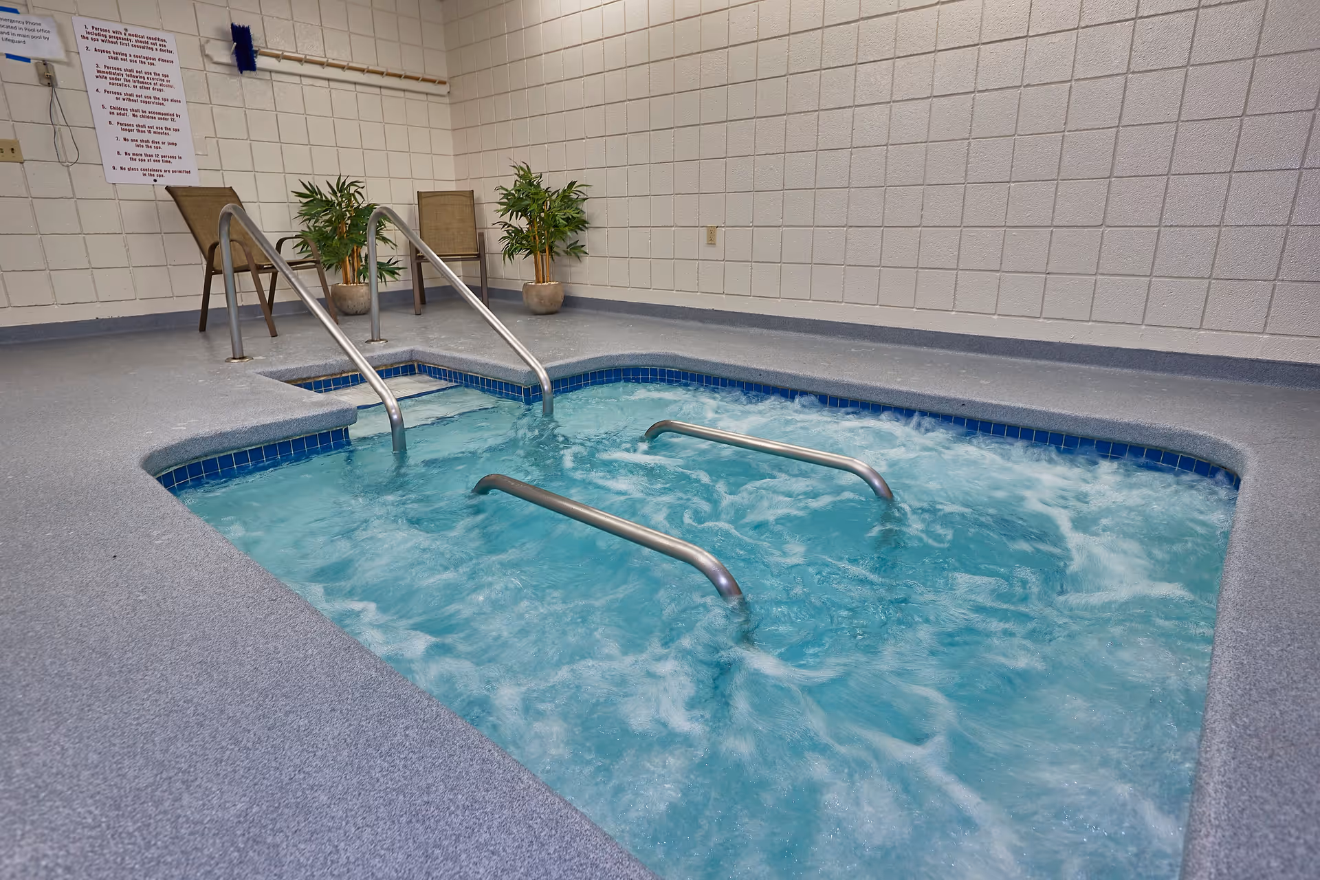 Indoor bubbling whirlpool spa with metal handrails, two chairs and potted plants against a tiled wall.